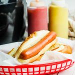 Red basket with hot dog in front of bottles of ketchup and mustard