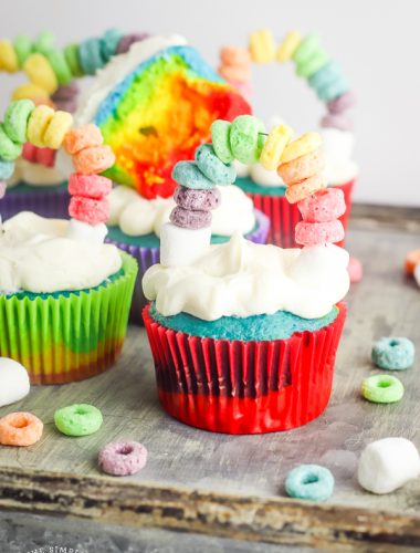 Rainbow cupcakes with rainbow cake topper sitting on cutting board