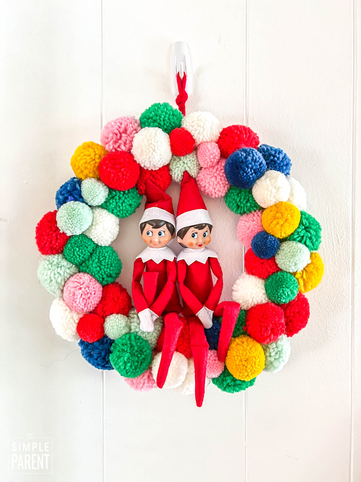 Two Christmas elves sitting in multi colored pom pom wreath